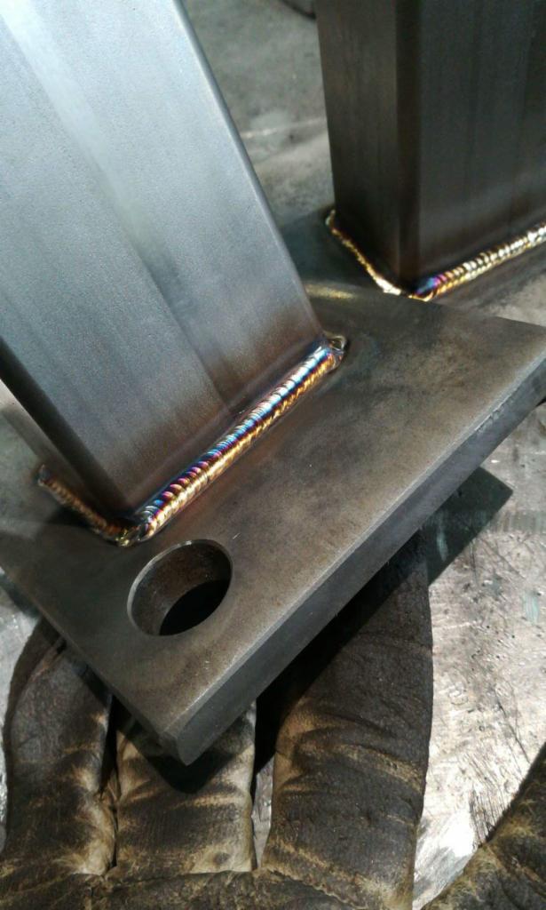 Metalowa konstrukcja spawana z widocznymi tęczowymi przebarwieniami spoiny, obok leżą robocze rękawice.