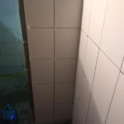 Świeżo ułożone, jasne, kwadratowe płytki ścienne z plastikowymi krzyżykami dystansowymi, widoczny fragment ściany przygotowanej pod płytki oraz urządzenie na podłodze.