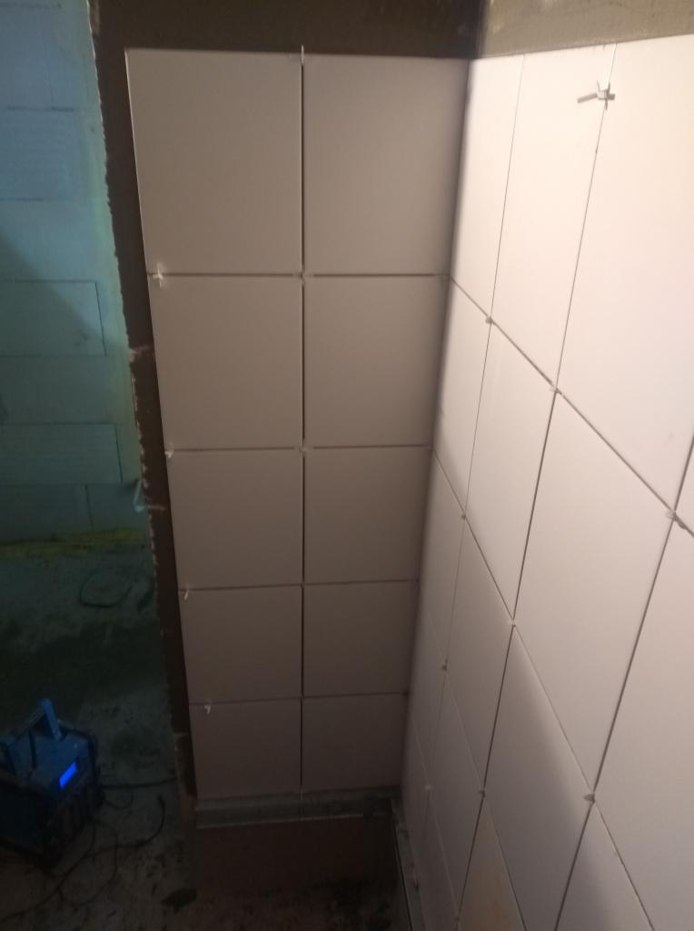 Świeżo ułożone, jasne, kwadratowe płytki ścienne z plastikowymi krzyżykami dystansowymi, widoczny fragment ściany przygotowanej pod płytki oraz urządzenie na podłodze.