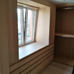 Jasna garderoba z drewna z wnęką okienną i szufladami, widok na zewnątrz przez uchylone okno, minimalistyczny design.