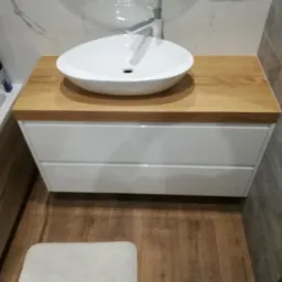 Umywalka nablatowa w kształcie łzy na drewnianym blacie z białymi szufladami, widoczna bateria i fragment wanny.