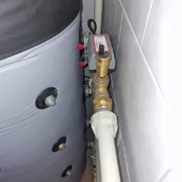 Szary zbiornik akumulacyjny ciepła z widocznymi przyłączami hydraulicznymi i zaworem Honeywell, zamontowany przy ścianie z szarych płytek.