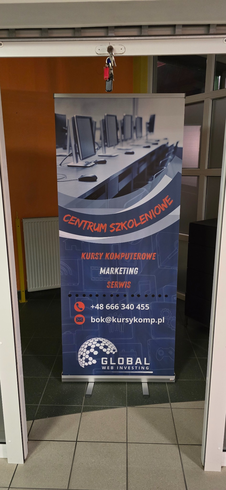 Roll-up reklamowy z informacją o centrum szkoleniowym oferującym kursy komputerowe i marketingowe, umieszczony w przejściu, z kluczami wiszącymi nad nim.