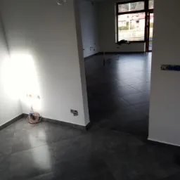 Wnętrze w trakcie prac wykończeniowych: świeżo położona ciemnoszara podłoga z płytek, gładkie białe ściany z widocznymi puszkami elektrycznymi i zwisającym przewodem, widok na kolejne pomieszczenie...