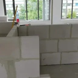 Wznoszenie ścian działowych z białych bloczków w surowym wnętrzu, widoczne narzędzia: poziomica, młotek, szpachelka, okna z widokiem na zieleń.