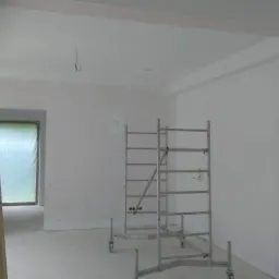 Wnętrze w trakcie malowania na biało, widoczne rusztowania, drabina malarska oraz fragment sufitu z listwą dekoracyjną.