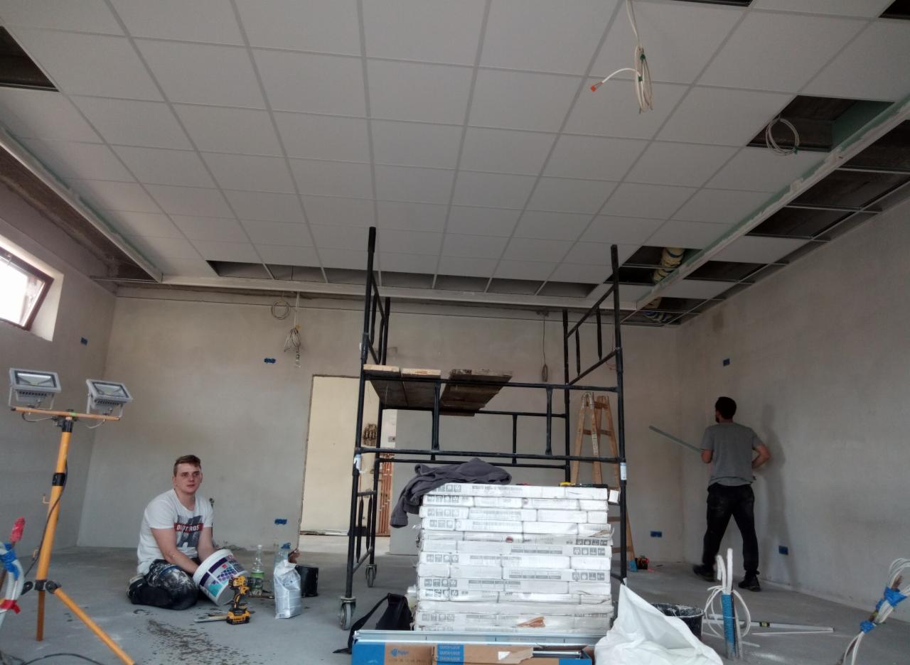 Wnętrze w trakcie prac wykończeniowych: pracownik siedzi na podłodze obok wiadra z farbą i narzędzi, drugi maluje ścianę, widoczne rusztowanie, płyty sufitowe i reflektory na statywie.