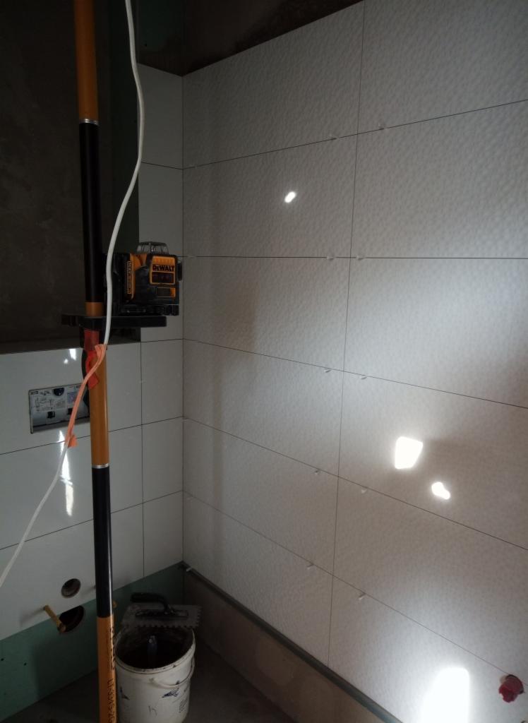 Ściana łazienki w trakcie układania jasnych, kwadratowych płytek z delikatną strukturą, poziomowanie laserowe, widoczny kubeł z zaprawą i szpachelka, etap prac wykończeniowych.