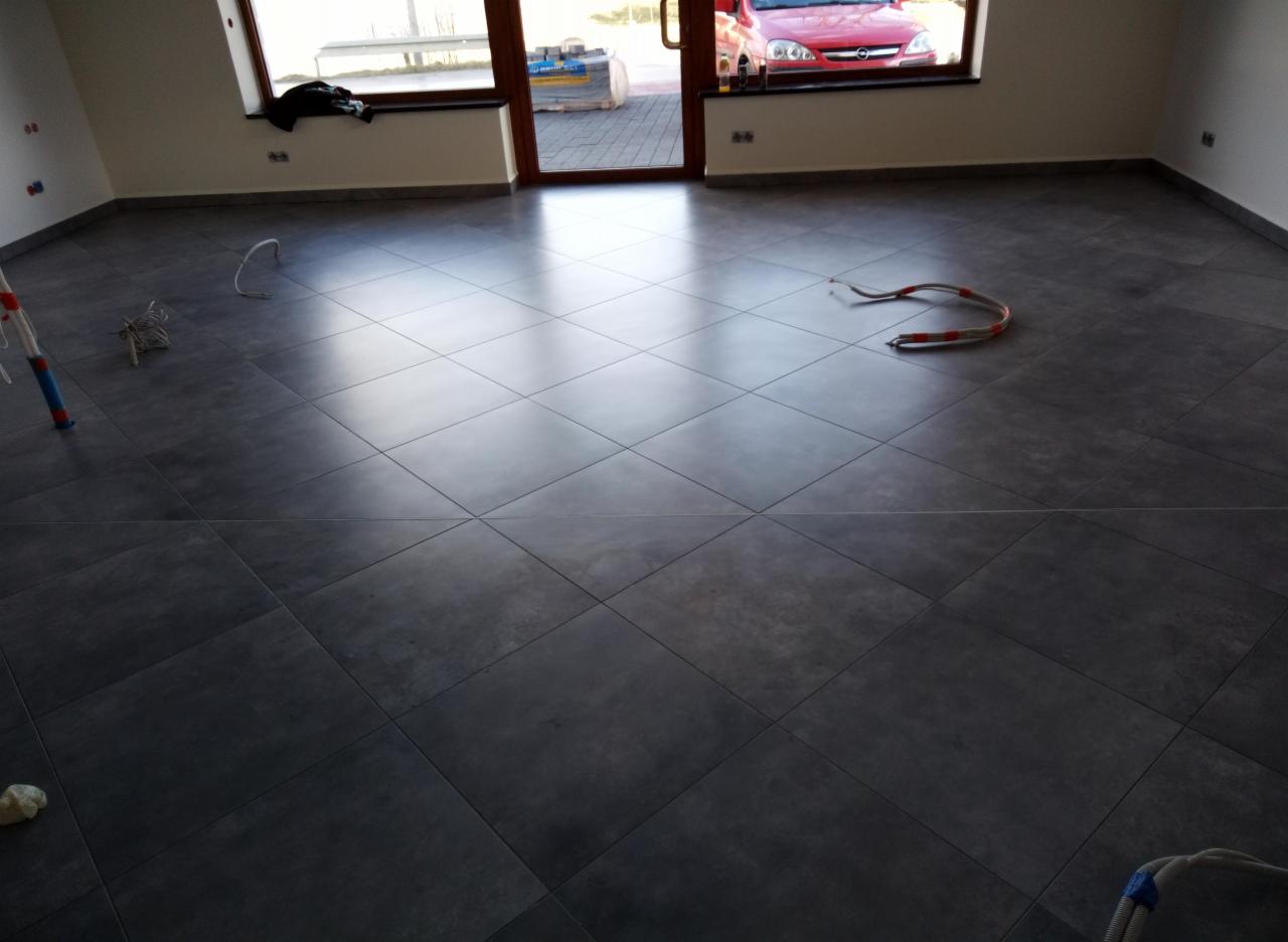 Nowo ułożona, ciemnoszara podłoga z płytek ceramicznych w przestronnym, jasnym pomieszczeniu z dużym oknem i drzwiami balkonowymi, przez które widać zaparkowany czerwony samochód.