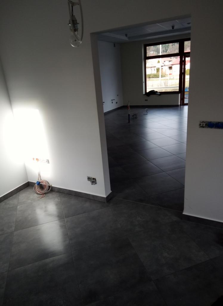 Wnętrze w trakcie prac wykończeniowych: świeżo położona ciemnoszara podłoga z płytek, gładkie białe ściany z widocznymi puszkami elektrycznymi i zwisającym przewodem, widok na kolejne pomieszczenie...
