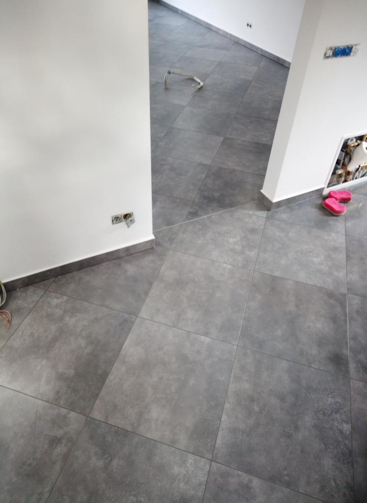 Ułożona szara, matowa podłoga z płytek ceramicznych w pomieszczeniu z białymi ścianami i wystającymi przewodami elektrycznymi. W tle widoczna rurka do instalacji wodnej oraz różowe klapki przy...