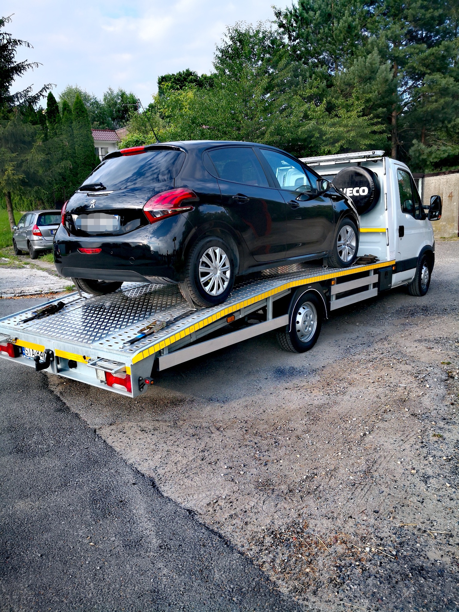 Czarny samochód osobowy marki Peugeot 208 zabezpieczony pasami na platformie lawety Iveco w trakcie transportu; widoczne tablice rejestracyjne lawety.
