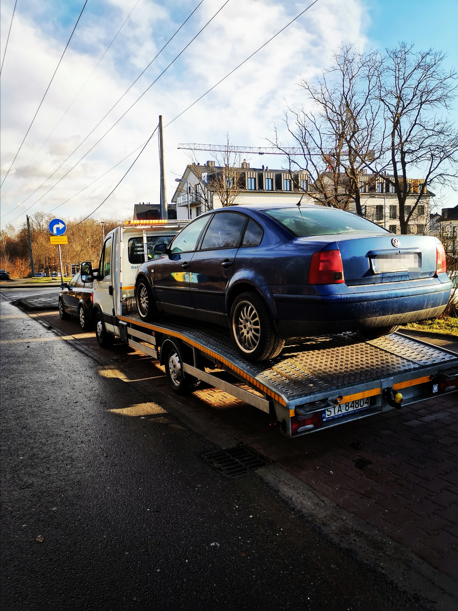 Niebieski samochód osobowy marki Volkswagen Passat przewożony na platformie lawety w słoneczny dzień na tle miejskiego krajobrazu z budynkami i drzewami.