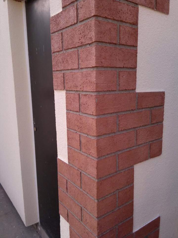 Fragment ocieplonej elewacji budynku z dekoracyjną cegłą klinkierową w narożniku, widoczna czarna stolarka drzwiowa.