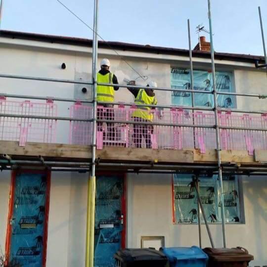 Prace na rusztowaniu przy ocieplaniu elewacji budynku w Hull, widoczne zabezpieczone okna i drzwi folią ochronną, pracownicy w kamizelkach odblaskowych. Częściowo ocieplona ściana budynku.