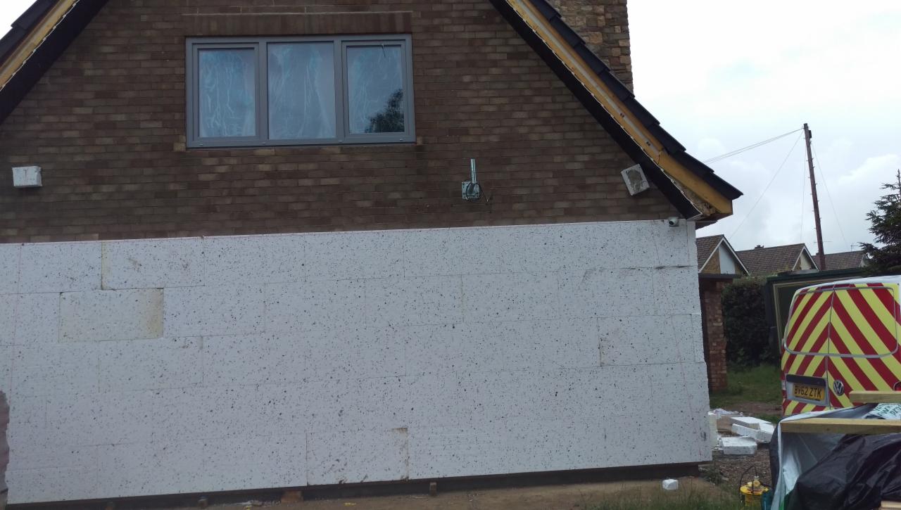 Częściowo ocieplona ściana domu jednorodzinnego z cegły, z widocznymi białymi płytami izolacyjnymi EPS, szara stolarka okienna, oraz zaparkowany samochód dostawczy z żółto-czerwonymi pasami.