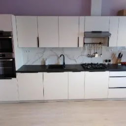 Nowoczesna, biała zabudowa kuchenna z czarnymi, minimalistycznymi uchwytami, czarnym blatem i marmurkowym panelem nad blatem, widok na kuchenkę gazową i piekarnik w zabudowie.