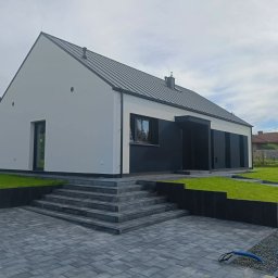 Nowy Dom s.c. - Nowoczesny dom jednorodzinny z elewacją w kontrastujących kolorach bieli i ciemnego grafitu, z geometrycznymi schodami z kostki brukowej i zadbanym trawnikiem.