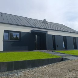 Nowy Dom s.c. - Nowoczesny dom jednorodzinny z kontrastową elewacją w odcieniach grafitu i jasnego beżu, z ciemnym dachem panelowym i minimalistycznym wejściem.