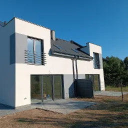 Nowoczesny dom jednorodzinny z jasną elewacją, szarymi akcentami i dużymi oknami, widok z zewnątrz na ukończoną elewację i częściowo wyłożony kostką taras, błękitne niebo w tle.