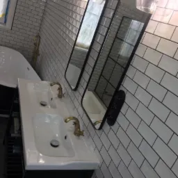 Wykończona łazienka z białą, prostokątną glazurą na ścianach, podwójną umywalką z czarną szafką i złotymi kranami, owalną wanną oraz dwoma wysokimi lustrami w czarnych ramach i lampami w stylu retro.