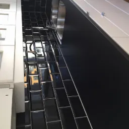 Błyszczące, czarne płytki ułożone w cegiełkę na ścianie nad blatem kuchennym, sąsiadujące z białymi szafkami z przeszklonymi frontami.