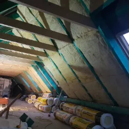 Izolacja poddasza wełną mineralną Isover Super-Mata, widoczne krokwie, kontrłaty, łaty, okno dachowe i rolki materiału izolacyjnego na podłodze