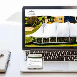 Laptop wyświetla stronę internetową firmy Tentrum prezentującą biały namiot imprezowy na tle zielonego trawnika i budynków. Smartfon leży na klawiaturze, pokazując mobilną wersję strony 'Hale...