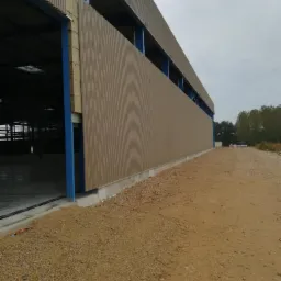 Budynek hali w trakcie budowy, widoczne brązowe panele elewacyjne z pionowymi żłobieniami oraz niebieska konstrukcja stalowa, surowy teren wokół.