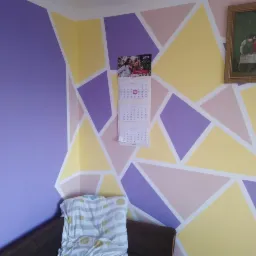Ściana w pokoju pomalowana w geometryczne wzory w odcieniach fioletu, żółci i różu, z wiszącym kalendarzem i obrazem w złotej ramie.