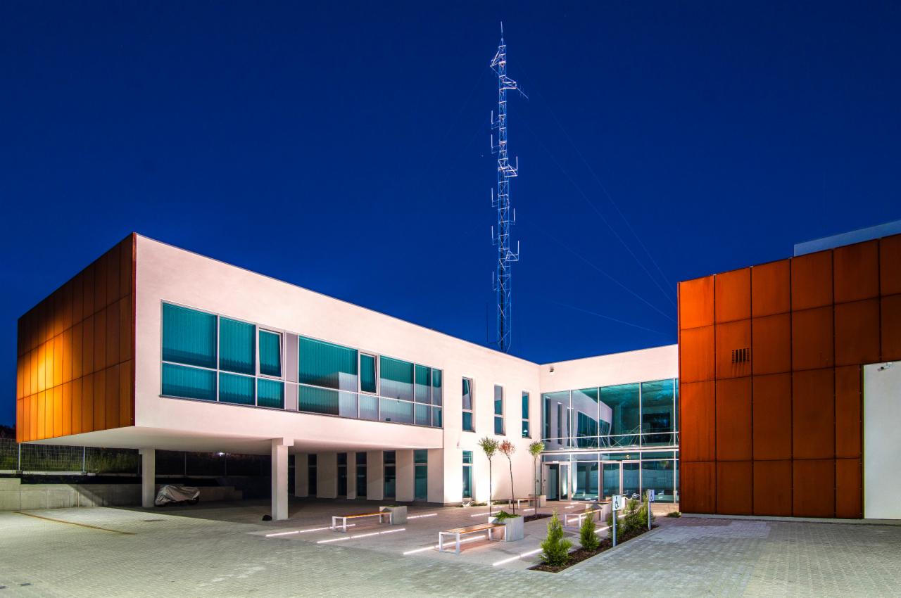 Nowoczesny budynek biurowy z elementami z blachy kortenowskiej, oświetlony w nocy, z wysoką wieżą telekomunikacyjną w tle.