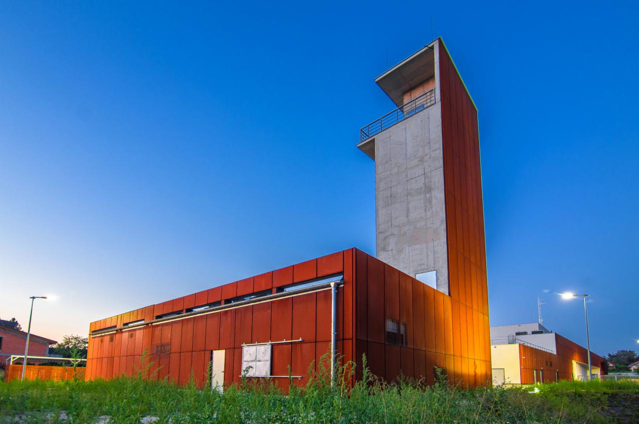 Nowoczesny budynek straży pożarnej z wysoką, betonową wieżą obserwacyjną obłożoną panelami w kolorze rdzy, oświetlony ciepłym światłem na tle błękitnego nieba o zmierzchu.