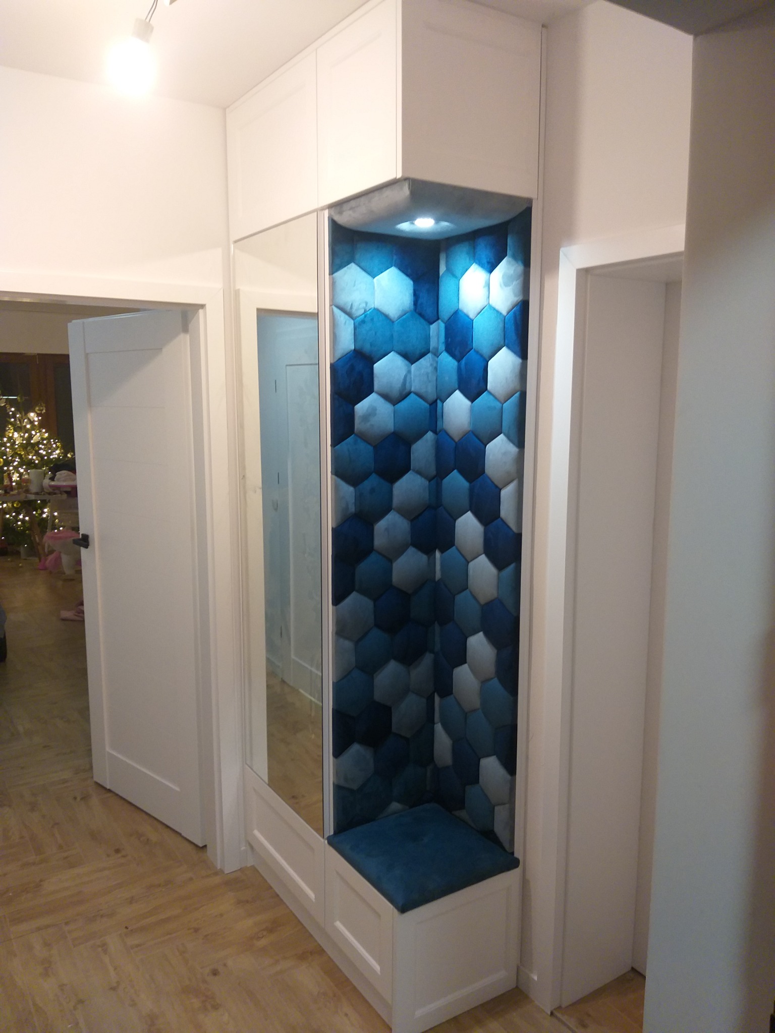 Wnęka z siedziskiem obita tapicerowanymi panelami w kształcie niebieskich i szarych sześciokątów, wbudowana w białą szafę z lustrem w przedpokoju.