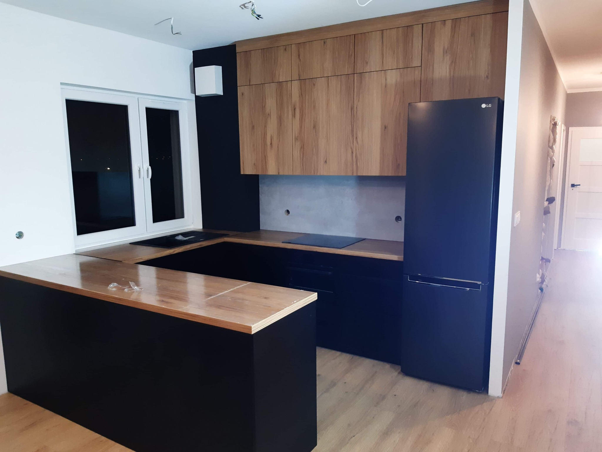 Nowoczesna kuchnia z czarnymi szafkami dolnymi i drewnianymi górnymi, wyspą z drewnianym blatem i czarną lodówką LG.