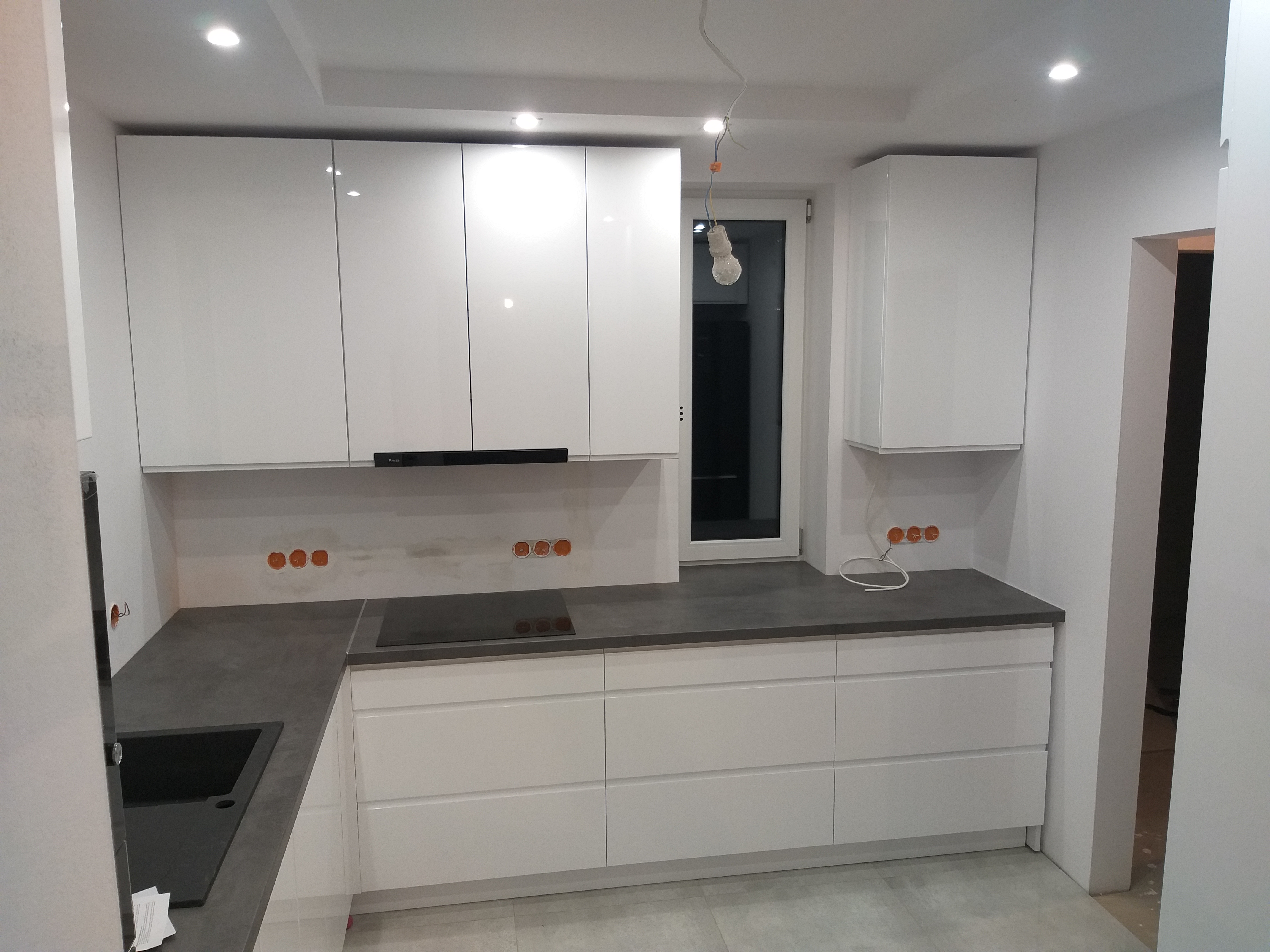 Nowoczesna kuchnia z białymi, lakierowanymi frontami szafek, szarym blatem i czarnym zlewem, jeszcze w trakcie montażu z widocznymi puszkami elektrycznymi i wiszącą żarówką.