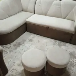 Beżowy narożnik tapicerowany z pufami na betonowej podłodze, widoczne przeszycia na oparciu w kształcie muszli.