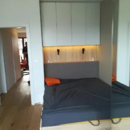 Wnęka sypialna z szarą tapicerowaną ścianą, oświetleniem LED i szafkami wiszącymi w jasnym odcieniu szarości, widok z boku z fragmentem podłogi z jasnego drewna.