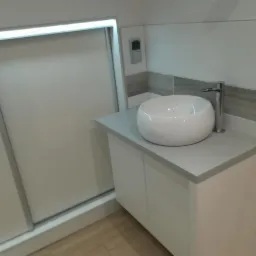 Nowoczesna łazienka z białą, okrągłą umywalką nablatową i minimalistyczną baterią, umieszczona na szafce z białymi frontami, obok przesuwanych drzwi z oświetleniem LED i szarymi akcentami na ścianie.