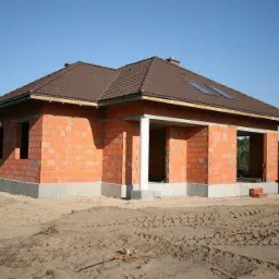 Surowy, niewykończony dom jednorodzinny z czerwonej cegły, z brązowym dachem i widocznym betonowym fundamentem, w otoczeniu piaszczystego terenu i drzew iglastych.