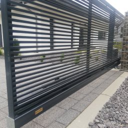 Dariusz Stepa METAL-HOUSE - Nowoczesna przesuwna brama wjazdowa z poziomych profili stalowych, widoczna z boku, z gabionowym ogrodzeniem w tle i kostką brukową na podjeździe.