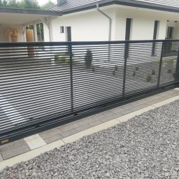 Dariusz Stepa METAL-HOUSE - Nowoczesna, pozioma brama wjazdowa przesuwna w kolorze czarnym, z wypełnieniem z poziomych profili, zamontowana w Żarach, z widoczną konstrukcją gabionową po prawej stronie i minimalistycznym domem...