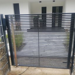 Dariusz Stepa METAL-HOUSE - Nowoczesna brama wjazdowa z poziomych, czarnych profili, osadzona na słupkach z gabionów wypełnionych kamieniami, na tle białego budynku z ciemnymi oknami.