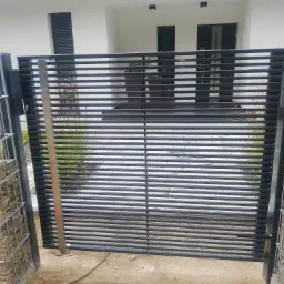 Nowoczesna brama wjazdowa z poziomych, czarnych profili, osadzona na słupkach z gabionów wypełnionych kamieniami, na tle białego budynku z ciemnymi oknami.