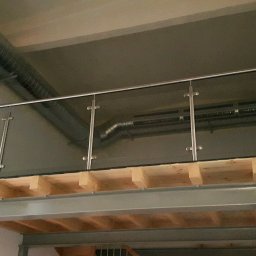Dariusz Stepa METAL-HOUSE - Szklana balustrada z nierdzewnymi słupkami na antresoli w industrialnym wnętrzu, widoczne drewniane belki konstrukcyjne i metalowe rury wentylacyjne pod sufitem.