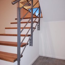 Dariusz Stepa METAL-HOUSE - Metalowa balustrada schodowa w kolorze szarym, z drewnianymi stopniami, zamocowana do białej ściany, widok z dołu na górę schodów.