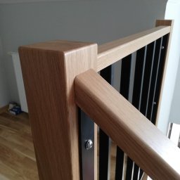 Dariusz Stepa METAL-HOUSE - Fragment balustrady schodowej z drewnianymi elementami w odcieniach beżu i czarnymi, pionowymi słupkami, mocowanymi śrubami, widok z góry na podłogę.