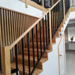 Drewniane schody z czarnymi metalowymi balustradami w nowoczesnym wnętrzu, widoczne elementy wykończenia z jasnego drewna i białe ściany.