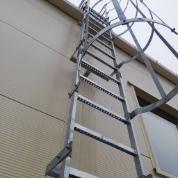 Dariusz Stepa METAL-HOUSE - Zewnętrzna metalowa drabina ewakuacyjna z koszem ochronnym przymocowana do ściany budynku z blachy trapezowej, widoczna z dołu pod kątem.