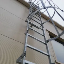 Zewnętrzna metalowa drabina ewakuacyjna z koszem ochronnym przymocowana do ściany budynku z blachy trapezowej, widoczna z dołu pod kątem.