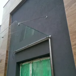 Szklany daszek nad wejściem do budynku, wsparty na metalowych wspornikach, z widocznymi kroplami deszczu na szkle i zabezpieczonymi folią oknami poniżej.